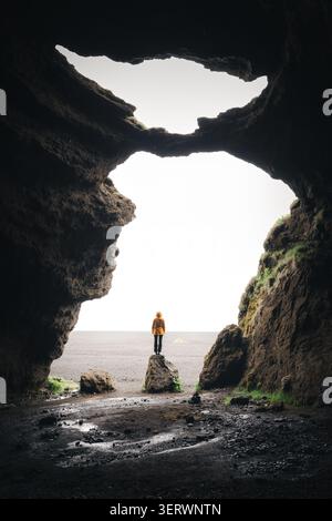 Blick auf eine Person in einer gelben Jacke, die auf einem Felsen in einer dunklen vulkanischen Höhle Yoda Caveopening steht und auf eine helle, bedeckte Landschaft blickt Stockfoto