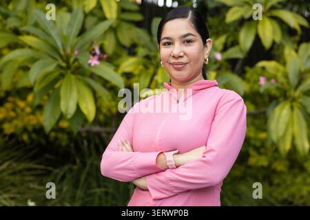 Frau, die mit überkreuzten Armen im Garten zwischen breiten Blättern steht, rosa Reißverschluss und Smartwatch trägt Stockfoto