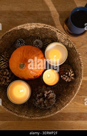 Runder Korb mit kleinem orangefarbenen Kürbis, brennenden Glaskerzen, Tannenzapfen auf Holztisch Stockfoto