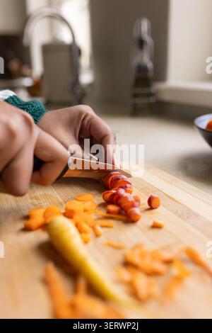 Erwachsene weibliche grüne Strickärmel schneiden dreifarbige Karotten mit Kochmesser auf Holzbrett am Waschbecken Stockfoto