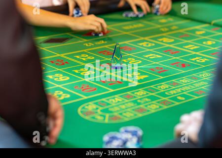 Eine Gruppe von Leuten versammelt sich um einen Roulette-Tisch in einem Kasino. Sie platzieren Wetten und beobachten, wie der Ball dreht. Die Atmosphäre ist lebhaft, da Chips gestapelt sind. Stockfoto