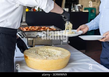 Ein Koch serviert Pasta von einem großen Käserad bis zu einem Gast bei einer Catering-Veranstaltung. Im Hintergrund stehen verschiedene Gerichte zur Verfügung. Gäste sind en Stockfoto
