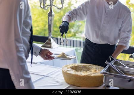 Bei einer Veranstaltung im Freien serviert ein Kellner Käsepasta. Die Umgebung hat Tische mit weißen Tüchern und grüne Bäume im Hintergrund. Stockfoto