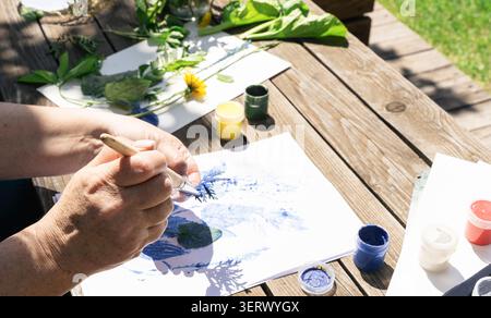 Künstlerin, die im Freien botanische Kunst kreiert und mit einem Pinsel blaue Farbe auf ein Blatt aufträgt, um natürliche Drucke zu erhalten Stockfoto