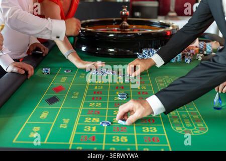 Eine Gruppe von Leuten versammelt sich um einen Roulette-Tisch in einem Kasino. Sie platzieren Wetten und beobachten, wie sich das Rad dreht. Die Getränke liegen auf dem Tisch und das Lachen ist groß Stockfoto