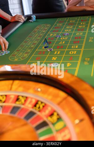 Die Leute versammeln sich abends an einem Roulette-Tisch in einem Kasino. Sie legen Chips auf Zahlen, während sie darauf warten, dass der Ball landet. Die Atmosphäre i Stockfoto
