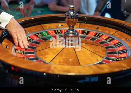 Die Leute versammeln sich um einen Roulette-Tisch in einem Casino-Setting. Eine Hand setzt eine Wette, während der Ball sich dreht. Die Atmosphäre ist lebendig, während die Spieler zusehen Stockfoto