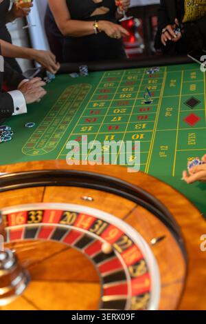 Eine Gruppe von Leuten versammelt sich um einen Roulette-Tisch in einem Kasino. Sie platzieren Wetten und beobachten das sich drehende Rad. Getränke gibt es in der Nähe, da sie ihre Gäste genießen Stockfoto