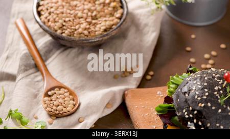 Dieser hochwinklige Speiseshot enthält einen modernen Burger mit einem schwarzen Brötchen mit Sesam-Belag, einen Holzlöffel und eine rustikale Schüssel mit trockenen Linsen, al Stockfoto