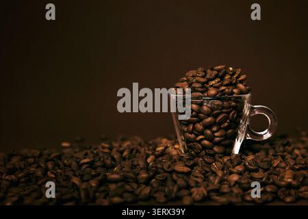 Geröstete Kaffeebohnen, die aus einer kleinen Tasse auf einem braunen Hintergrund herausfließen Stockfoto