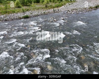 Der rauschende Fluss fließt über glatte Steine. Wasser wirbelt weiß und schaumig bei schneller Strömung. Die Gräser am Bankrand schwanken sanft im Wind. Die Steine am Fluss zeigen Stockfoto
