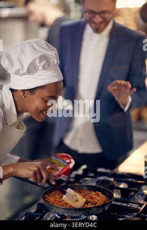 Kochen Sie in professioneller, moderner Küche, Restaurant-Konzept. Lächelnde Küchenchefin, die Essen in der Pfanne zubereitet. Stockfoto