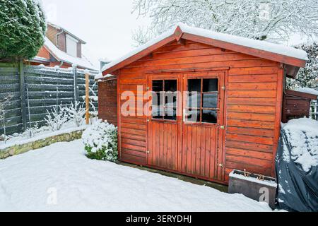 Hölzerner Gartenschuppen mit Schnee, warmen Holztönen, kontrastierendem weißem Dach, kleinen Fenstern und verschlossener Tür Stockfoto