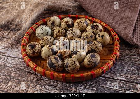 Großaufnahme von gesprenkelten Wachteleiern, angeordnet in einem runden Bambuskorb mit roten Bindungen, sitzend auf einer rustikalen Holzfläche mit getrocknetem Pampasgras ein braun Stockfoto