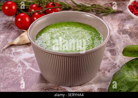 Leuchtend grünes, cremiges Suppenpüree in strukturierter grauer Schüssel, umgeben von frischen Kirschtomaten, Rosmarin, Knoblauch, Spinatblättern, roten Pfefferkörnern auf Rusti Stockfoto