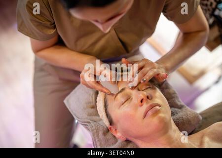 Kosmetikerin im Salon sanft schrubbt weibliche Klientelgesicht Haut mit Gua Sha Massage Steinwäscher, Gua Sha alte traditionelle chinesische Medizin Technik mit sanftem Hautkratzen mit Werkzeugen. Stockfoto