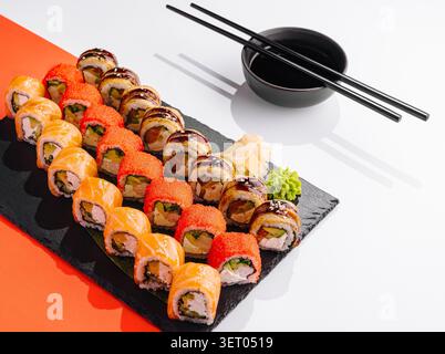 Verschiedene Sushi-Brötchen sind ordentlich auf einem schwarzen Teller angeordnet. Es gibt verschiedene Arten von Sushi, Saucen, Wasabi und Ingwer an der Seite. Stäbchen werden auf den Tisch gelegt. Stockfoto
