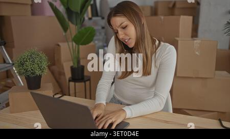 Junge Frau, die das Telefon am Ohr hält und auf einem Laptop in gestapelten Umzugskästen im Gebäude schreibt; Stress. Stockfoto