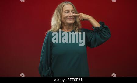 Ältere Frau mit blonden Haaren zeigt den Finger an die Nase, lächelt, trägt grüne Bluse vor rotem Studio-Hintergrund; verspieltes Selbstvertrauen. Stockfoto