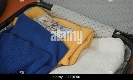 Frau, die einen Hartschalenkoffer packt, Hand mit blauen Nägeln mit Strohhut und sichtbaren Dollar auf dem Bett im Gebäude; Vorausschau. Stockfoto