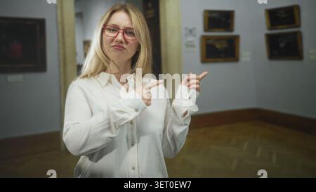 Frau in weißem Hemd mit roter Brille zeigt mit dem Finger auf gerahmte Gemälde an der Galeriewand im Museum, stehend und mit Händen gestickt, neutrale Ausdrücke Stockfoto