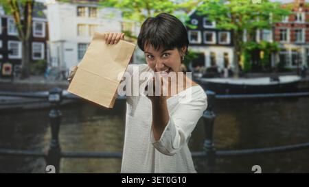 Die Frau zeigt mit dem Finger auf die Papiertüte auf der Kanalstraße, hält sie hoch und lächelt breit; Überraschung. Stockfoto