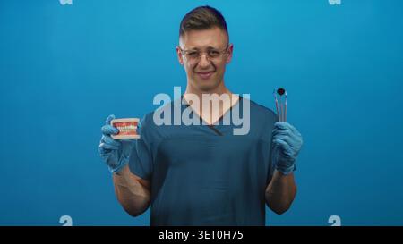 Männlicher Zahnarzt junger blonder in blauen Peelings und Handschuhen, die Zahnmodell und Spiegelwerkzeug mit Handschuhen halten, grimmig während Brille im Atelier; d Stockfoto