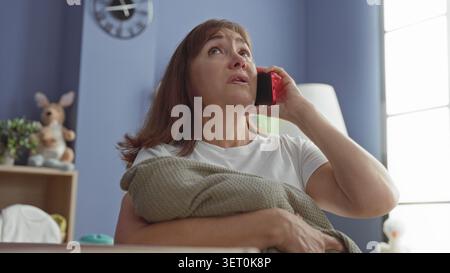 Die Frau hält das Smartphone an das Ohr und umarmt die Decke im Babyzimmer im Gebäude; Sorgen, Müdigkeit, Stress, überwältigt. Stockfoto