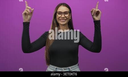 Die junge Frau zeigt beide Zeigefinger nach oben, trägt eine Brille und zeigt Finger und Gesicht in einem Studio; Selbstvertrauen feiern. Stockfoto
