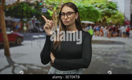 Junge Frau zeigt den Zeigefinger nach oben auf die Straße, Arme gefaltet, Brille und Jeans getragen; Vertrauensunterweisung. Stockfoto