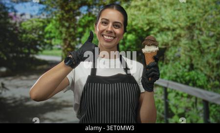 Frau auf Schürze, die Eisbecher hält und im Wald eine gute Geste macht, lächelt in die Kamera; fröhlicher Service. Stockfoto