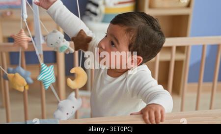 Der Junge greift nach einem hängenden Mobile, während er im Stehen steht und in einem Kinderzimmer die Gitterstange greift; Neugierde spielen. Stockfoto