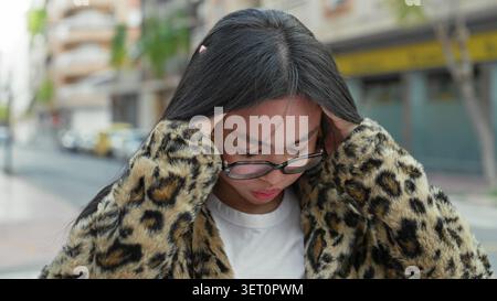 Frau mit Brille, die ihre Schläfen gegen Kopfschmerzen hält, Hände an Kopf auf einer Straße in Leopardenmantel; Stress, Konzentration. Stockfoto