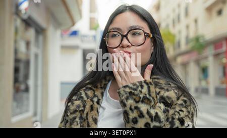 Frau, junge Latina, mit Brille, die Hand winkt auf einer Straße in der Nähe von Geschäften, trägt Leopardenmantel und lächelt breit; Selbstvertrauen. Stockfoto