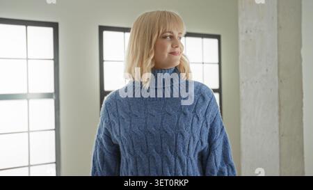 Frau in blauem Zopfstrick-Rollkragenpullover lächelt mit dem Kopf zur Seite gewendet, die Augen blicken aus großen Fenstern in hellem Studio; ruhige Reflexion. Stockfoto