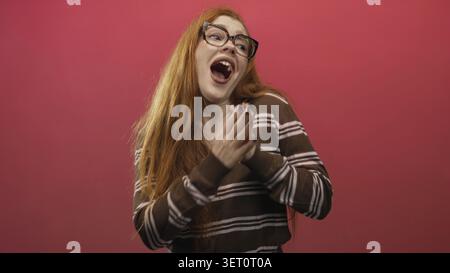 Rothaarige Frau, die Hände an die Brust schließt, lächelt mit geschlossenen Augen vor rosa Hintergrund in einem Studio; Freude. Stockfoto