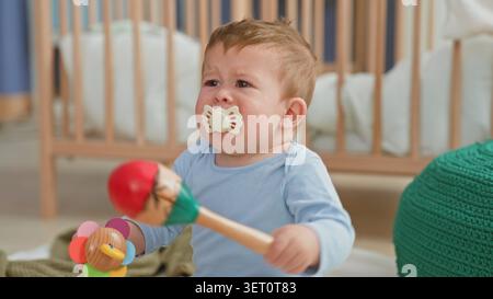 Kleinkind mit farbenfroher Rassel und Schnuller im Mund, der in einem Gebäude weint; Not sucht nach Komfort. Stockfoto