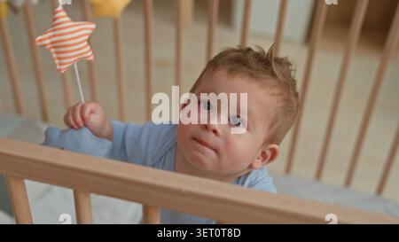 Kleinkind Junge hispanic weint, greift nach orange gestreiftem Sternenspielzeug und packende Gitterstange in hölzernem Kinderbett im Gebäude; Distress braucht Komfort. Stockfoto