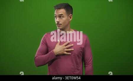 Mann mit rechter Hand auf der Brust und mit Handschnürlippen im Studio mit grünem Hintergrund, seitlichem Blick und zögerlicher Haltung, die Selbstzweifel und Überraschung suggeriert; V Stockfoto