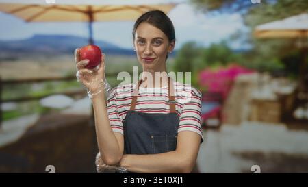Frau, die Apfel in Handschuhen auf einem Terrassengebäude vor dem Restaurant hält, trägt Schürze und gestreiftes Hemd, Arm gefaltet; Selbstvertrauen. Stockfoto