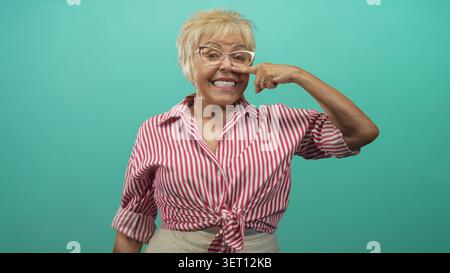 Eine ältere hispanische Frau mit Brille und einem gebundenen rot gestreiften Hemd zeigt den Zeigefinger auf ihre Nase, während sie vor einem türkisfarbenen Hintergrund in Studi lächelt Stockfoto