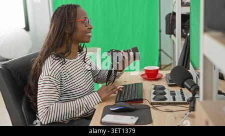 Frau hält Kamera und Typen auf der Tastatur in einem Studio mit grünem Bildschirm und roter Tasse auf dem Schreibtisch; fokussierte Kreativität. Stockfoto