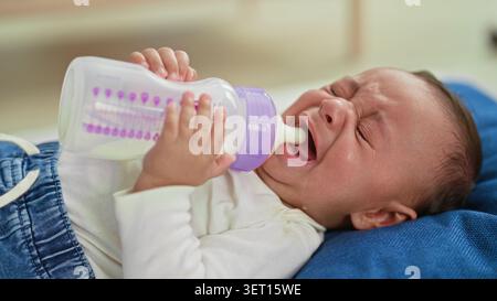 Hispanischer Junge weint, während er eine Milchflasche hält, eine kleine Hand am Brustwarzen hält und die Hand des Pflegepersonals beim Bauen hilft; Sestress Comfort Bonding. Stockfoto