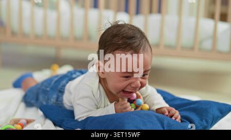 Junge weinend mit offenem Mund, der bunte Rassel auf blauem Kissen auf dem Bett im Gebäude hält; Traurigkeit Verletzlichkeit. Stockfoto