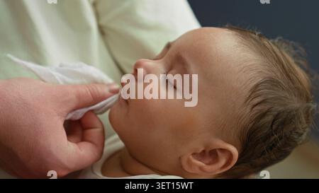 Hispanischer Junge, der auf dem Arm des Erwachsenen schläft, während die Hand das Gesicht sanft mit dem Tuch im Gebäude abwischt; sanfte Pflege beruhigt. Stockfoto