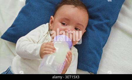 Hispanischer Junge, der weint und eine Milchflasche an den Mund hält, auf einem blauen Kissen in einer Futterszene im Studio, mit sichtbaren Tränen und geballter Hand; Hunge Stockfoto