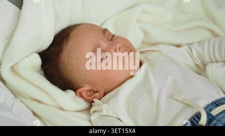 Hispanischer Kleinkind, der mit offenem Ohr und geschlossenen Augen auf einem weißen Bettbett im Gebäude schläft; friedliche Ruhe. Stockfoto