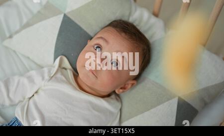 Der hispanische Junge, der in einem hölzernen Kinderbett auf einem gemusterten Kissen liegt, blickt mit weit offenen Augen und einem sichtbaren Gesicht auf die Kamera im Gebäude. Neugier Stockfoto
