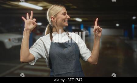Die blonde Frau im mittleren Alter mit Denim-Schürze macht mit der linken Hand ein OK-Schild und zeigt mit der rechten Hand den Zeigefinger nach oben in ein Parkhaus mit Auto Stockfoto