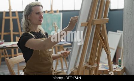 Die Malerin an der Staffelei reicht Hand mit dem Pinsel in Richtung Leinwand im Kunststudio, trägt braune Schürze und sitzt auf einem Stuhl; fokussierte Kreativität. Stockfoto